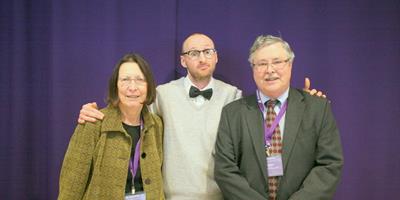 tony-and-joanne Three people posing together in front of a purple backdrop, two wearing conference name badges
