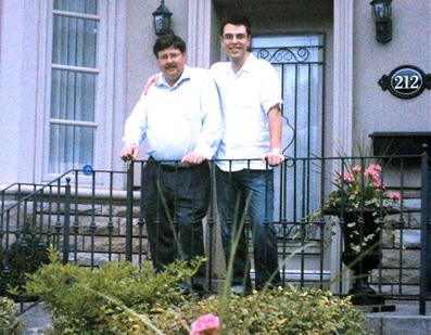 tony-and-john Two people standing side by side in front of a house entrance with flower planters