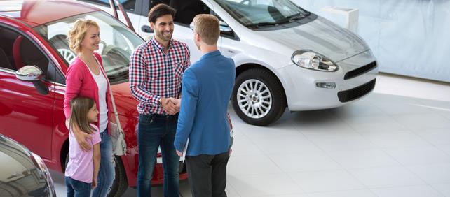 Salesperson greeting a family with two children beside a silver car in a car dealership showroom