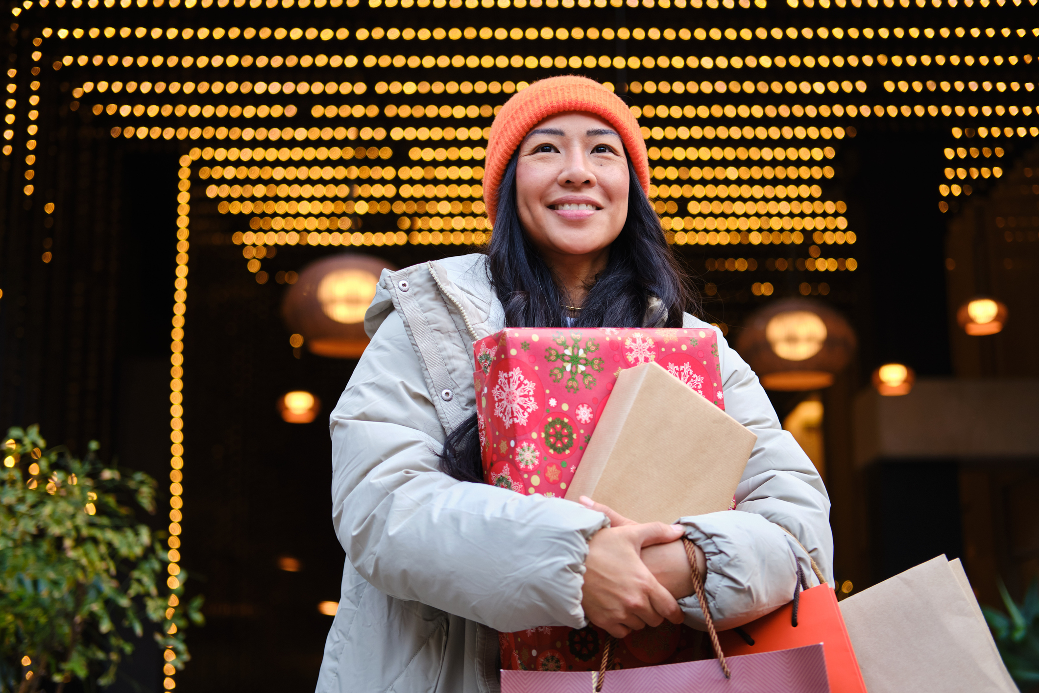 Smiling person in an orange toque holding wrapped holiday gifts and shopping bags under glowing lights