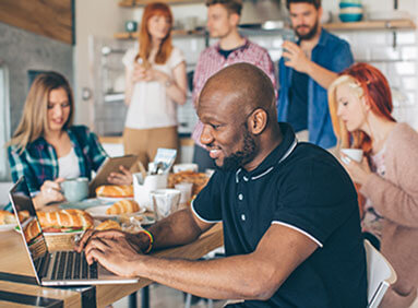 Person smiling while working on a laptop at a shared café table with people behind