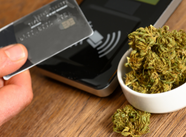 Hand tapping debit card on a machine with a small bowl of cannabis bud beside it