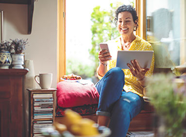 Smiling person sitting on the arm of a chair and holding a tablet in a bright living room
