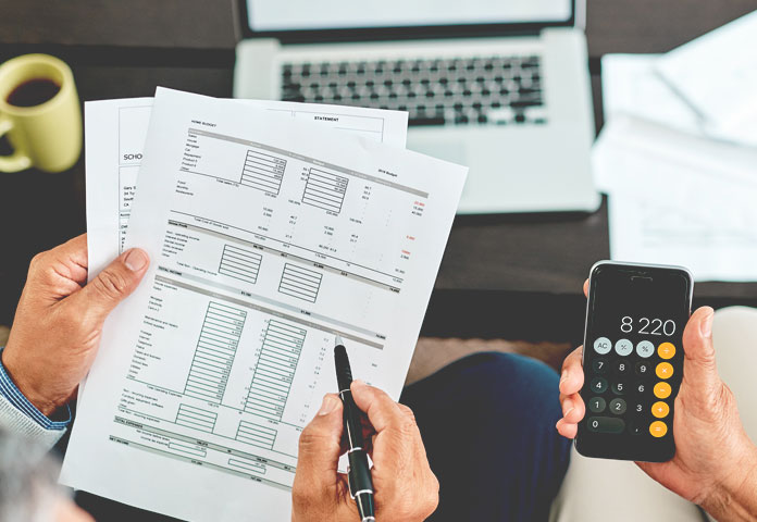 Hands holding financial documents and a phone calculator beside a laptop and coffee mug