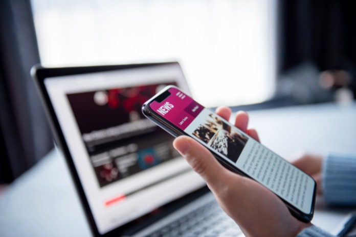 Hand holding a mobile phone with a news site on screen in front of a laptop, illustrating ClickScapes digital insights