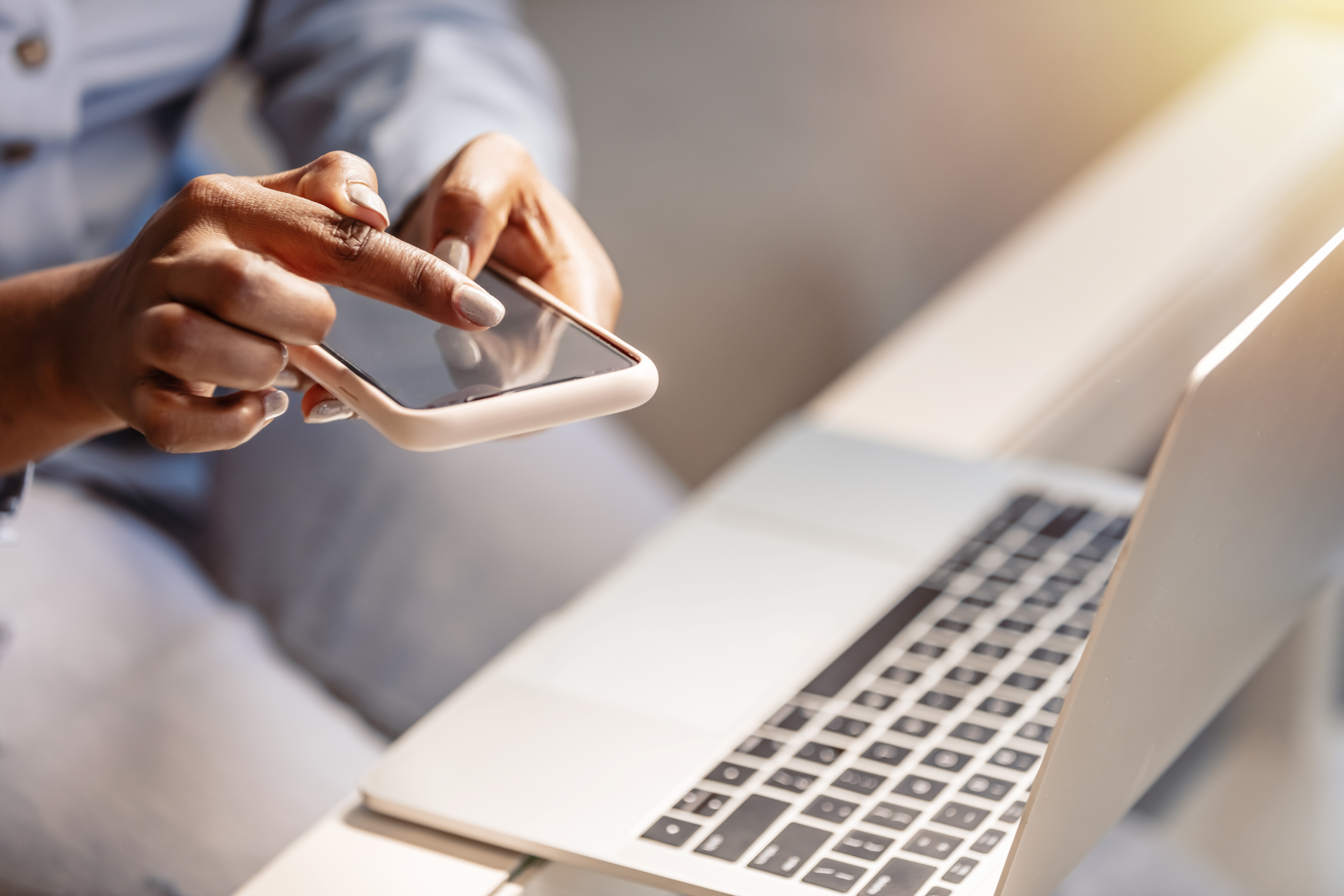 Hands typing on a cellphone with a laptop on a desk