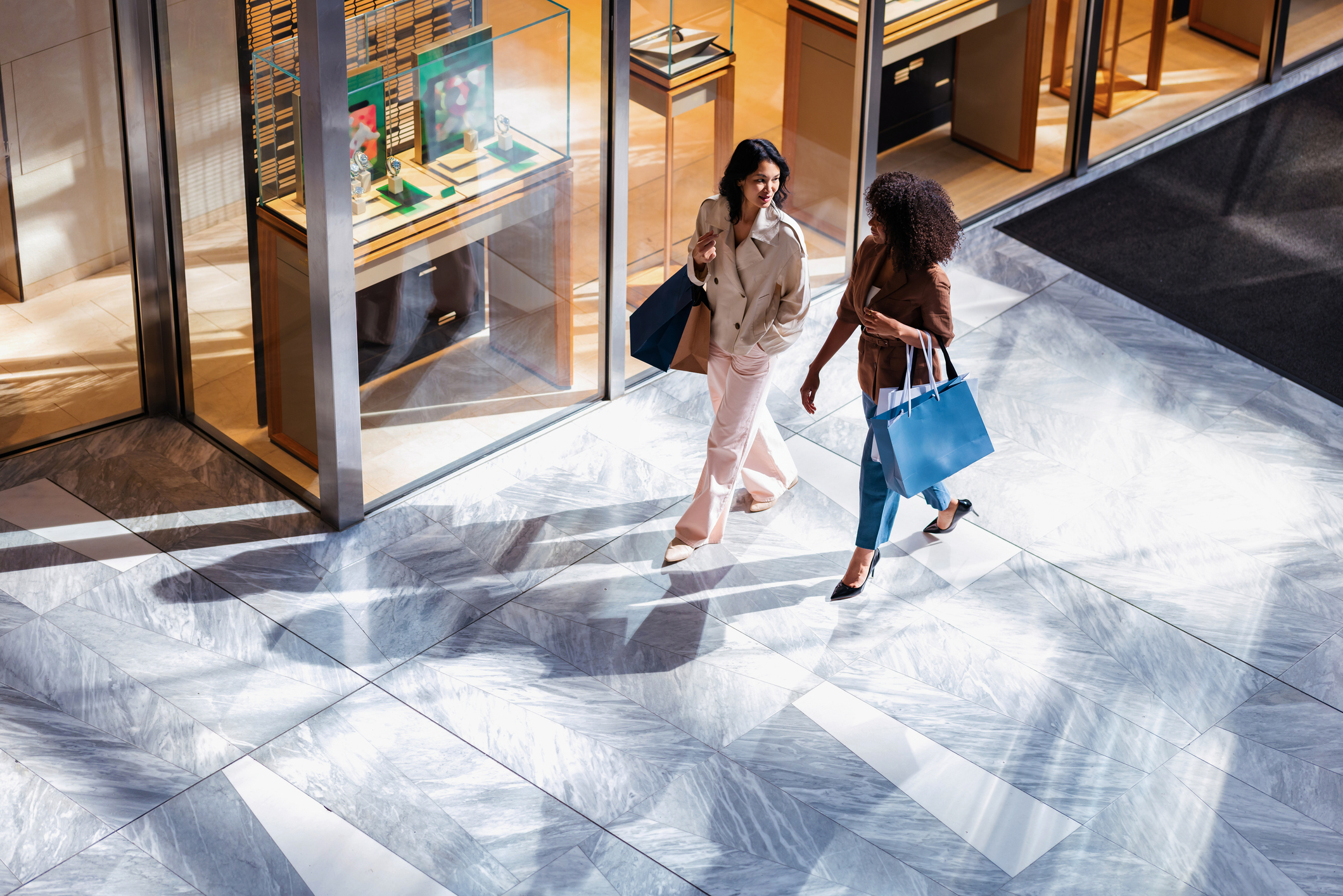 Two Friends Shopping In A Bright Modern Mall Carrying Bags And Chatting By Glass Storefronts