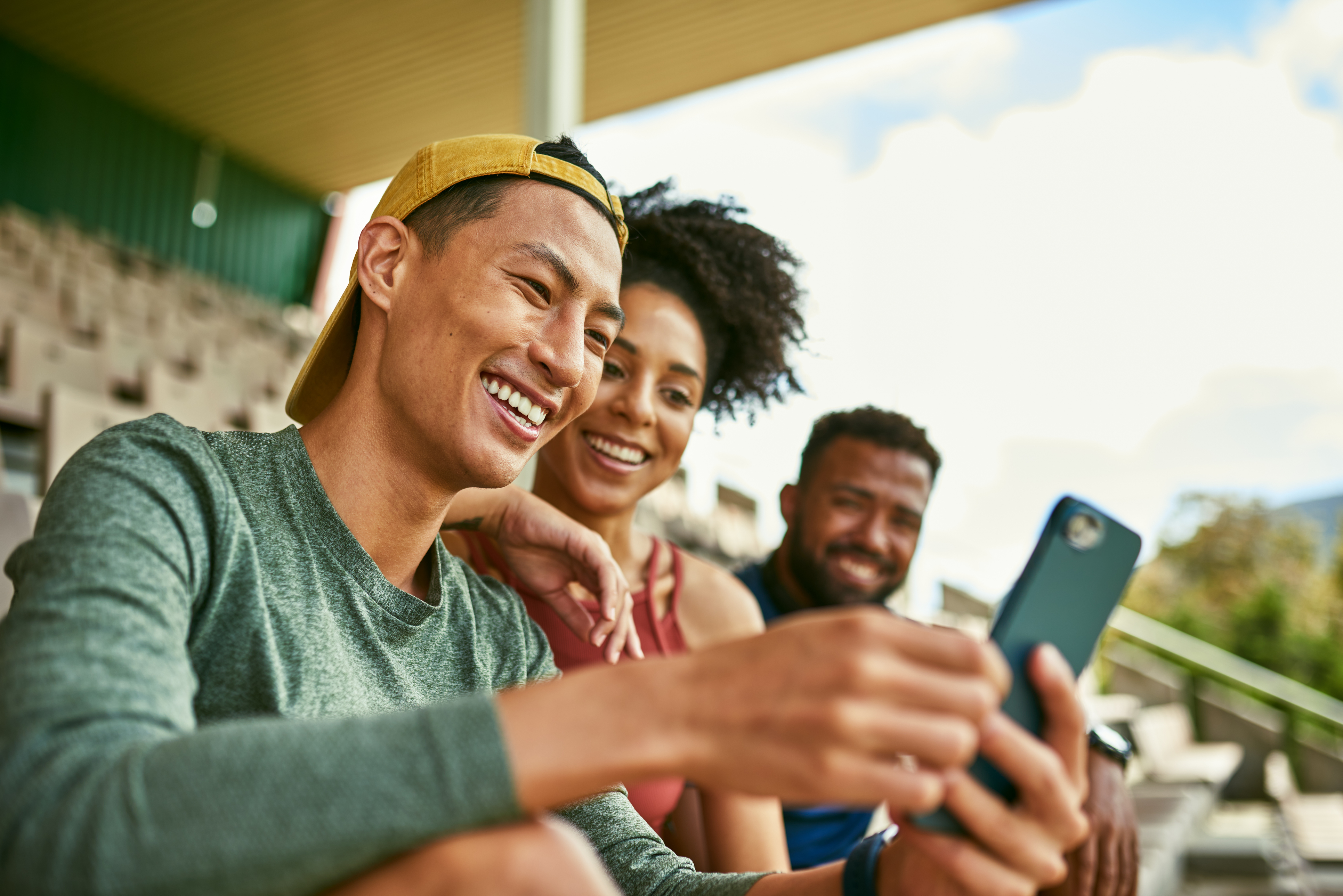 Young diverse group looking and laughing at website on mobile excitedly while sitting on stadium bleachers
